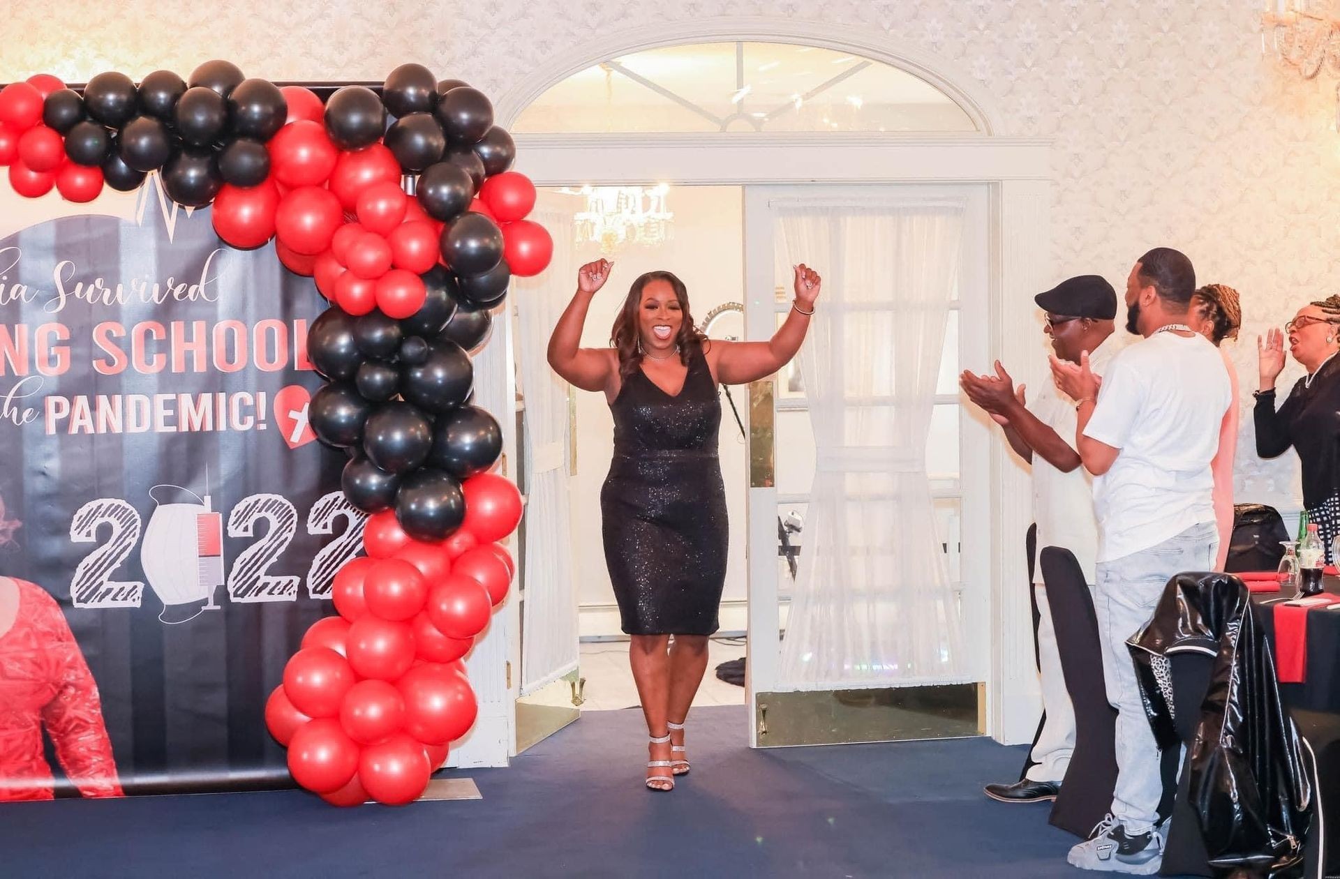 Woman celebrating in front of a balloon arch with people clapping during a party.