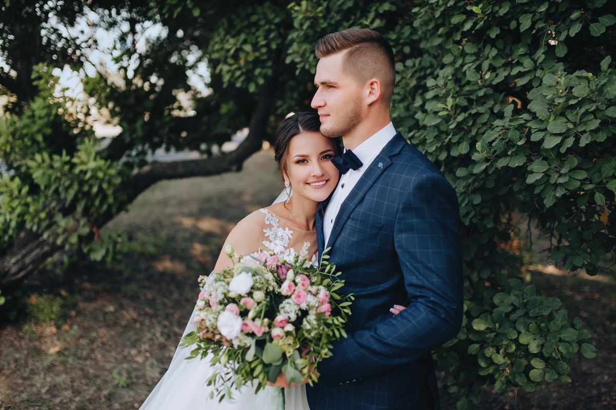 Beautiful newlyweds cuddle in a green garden with trees. Young bride and cute bride with a bouquet are standing in the park. Wedding photography. Stylish portrait.