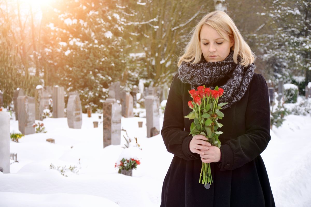 Woman in a snowy cemetery in winter with a bouquet of roses in deep mourning over death and loss